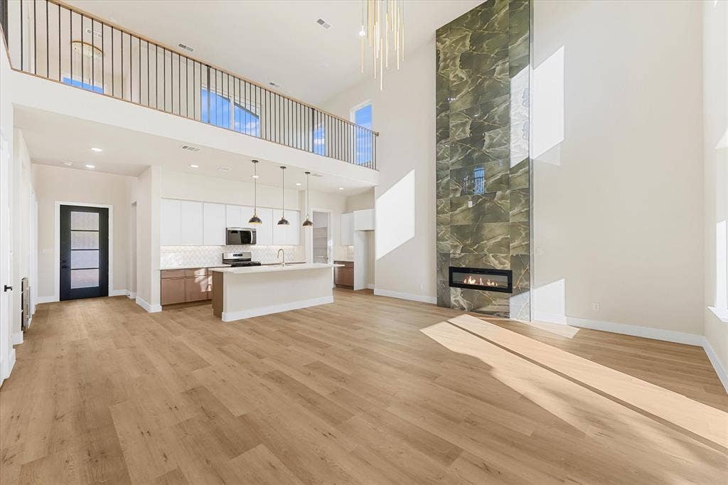Double-height ceiling, floor-to-ceiling stone-tiled fireplace, wood-finish flooring, island kitchen with white cabinetry, and an interior mezzanine with metal balusters