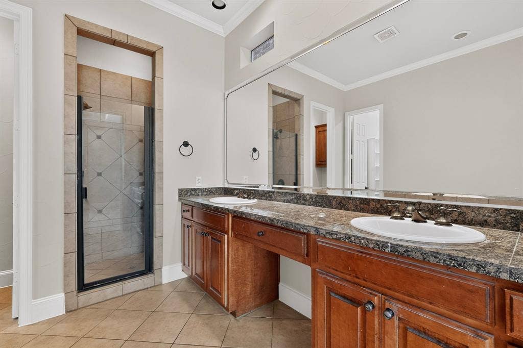 18359 Frankford Lakes Circle, Dallas, TX 75252 Double vanity with wood cabinetry, stone-finish countertops, dual white drop-in sinks, full-width wall mirror, and walk-in shower with tiled surround