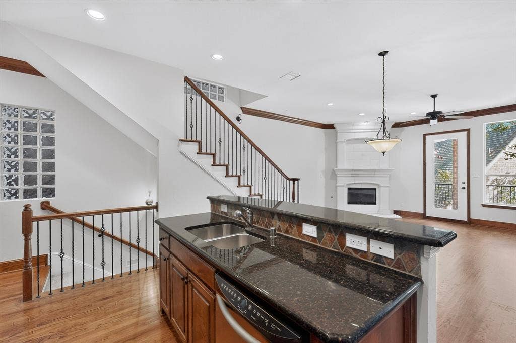 18359 Frankford Lakes Circle, Dallas, TX 75252 Kitchen featuring a dark stone countertop, undermount stainless steel sink, and wood cabinetry, with an interior staircase accented by decorative metal balusters, glass block accents, and a built-in fireplace