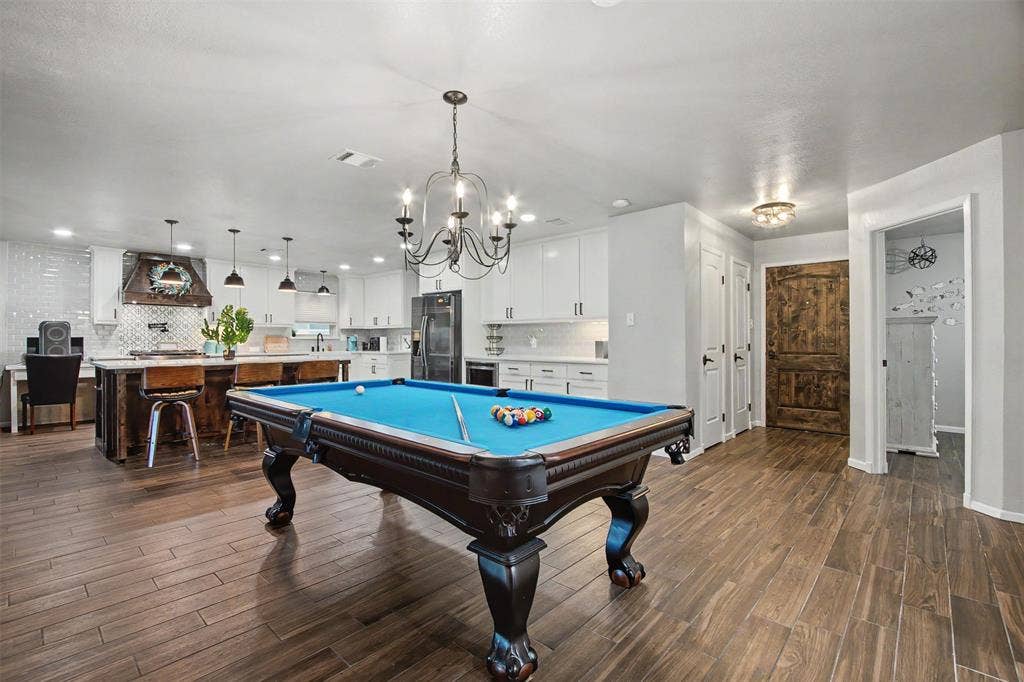 Open-concept space featuring wood-finish tile flooring, white shaker-style cabinetry, a central kitchen island with seating, a wrought-iron chandelier, and a rustic wood door