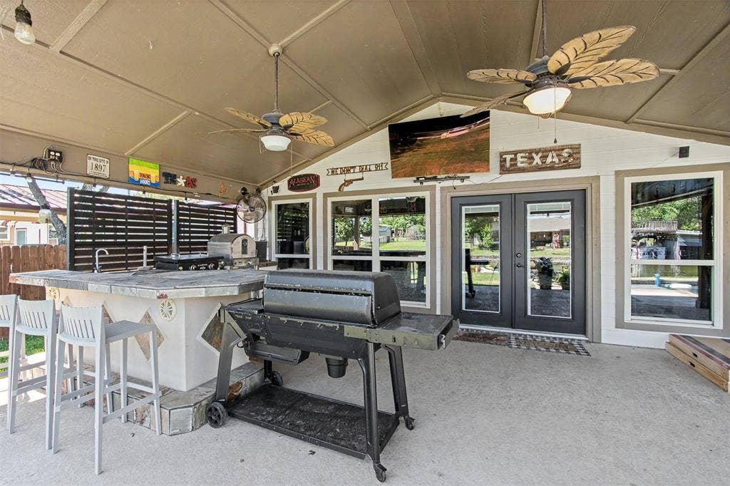 Covered patio featuring an outdoor kitchen with a stone-topped bar, dual ceiling fans, and double-door entry