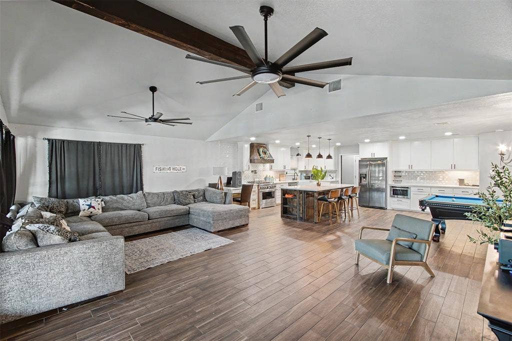Vaulted ceiling featuring exposed structural beams and dual multi-blade ceiling fans, wood-finish flooring, central kitchen island with bar seating, and white shaker-style cabinetry