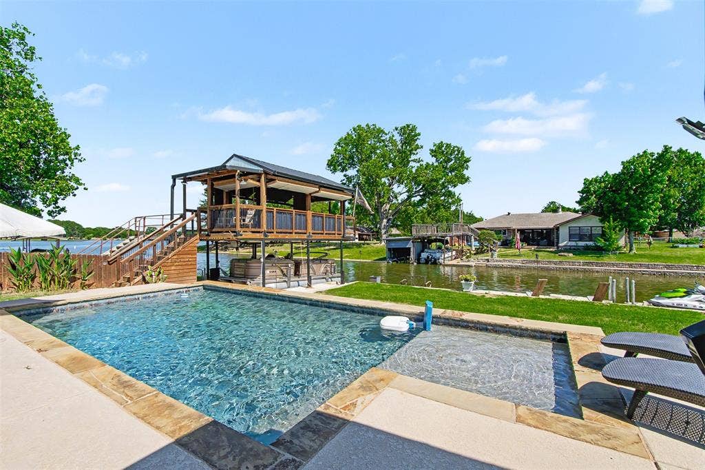 In-ground swimming pool featuring stone coping and an integrated sun shelf, elevated wooden boat dock with a covered upper deck, and manicured lawn