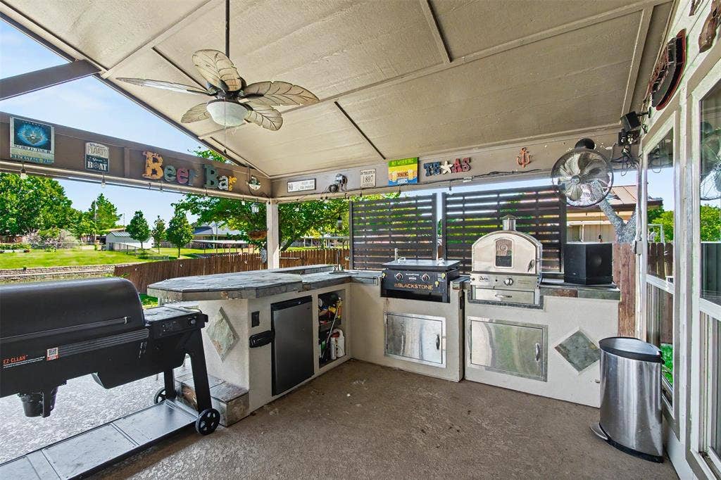 Covered outdoor kitchen featuring a concrete countertop, built-in stainless steel appliances, stacked stone accents, and a ceiling fan
