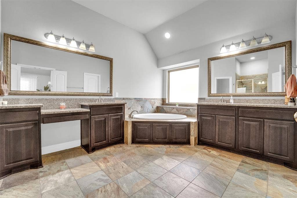 102 Champion Drive, Wylie, TX 75098 Bathroom featuring dual dark-wood cabinetry, stone-tiled flooring, drop-in soaking tub with stone surround, and dual wall-mounted vanity mirrors
