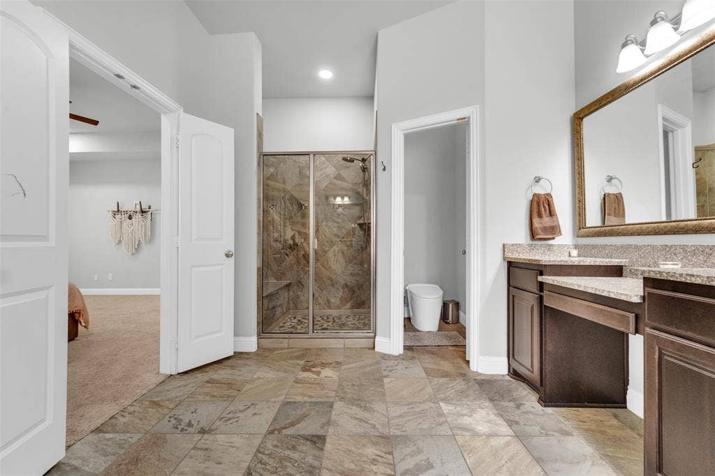 102 Champion Drive, Wylie, TX 75098 Bathroom featuring stone-tile flooring, walk-in shower with stone surround, dark-finish cabinetry, stone-style countertop, and a large wall-mounted mirror