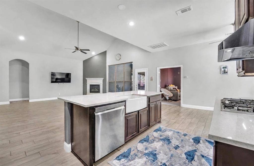 102 Champion Drive, Wylie, TX 75098 Open-concept kitchen featuring an island with a farmhouse sink, stainless steel appliances, stone-surface countertops, wood-finish flooring, and a fireplace with a decorative mantel