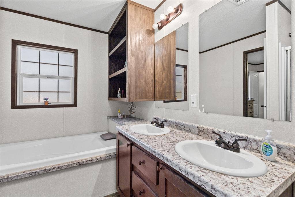 Bathroom featuring a dual-sink vanity with stone-patterned laminate countertops, wood-finish cabinetry, built-in shelving, and a soaking tub with matching surround