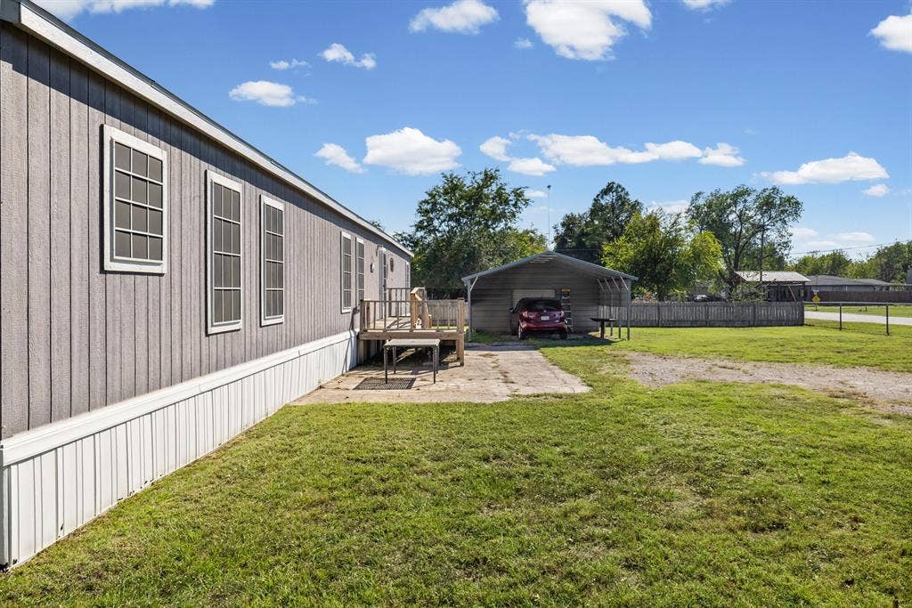 Exterior featuring vertical panel siding, a wood-deck entryway, a concrete patio, a metal-roofed carport, and fenced perimeter