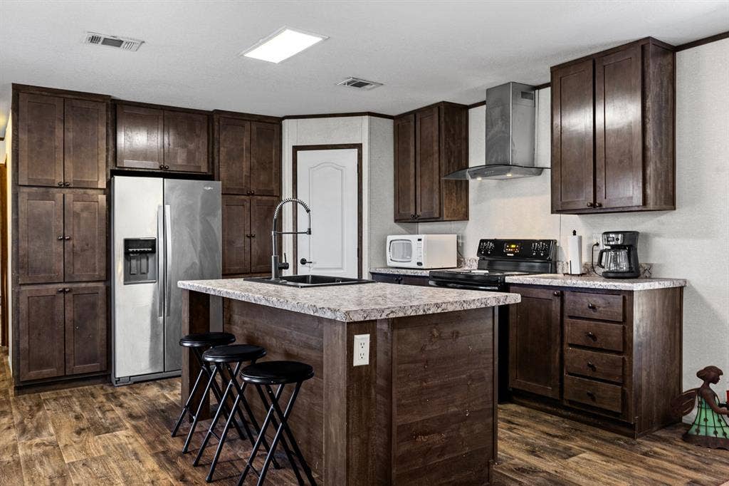 Kitchen featuring stained wood cabinetry, stone-patterned laminate countertops, a central island with a built-in sink, stainless steel kitchen appliances, and wood-finish flooring