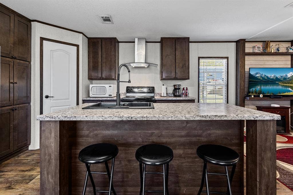 Kitchen featuring a central island with a stone-finish countertop, dark wood-tone cabinetry, a stainless steel chimney-style range hood, and a gooseneck faucet with an integrated sink, room for seating at the island.