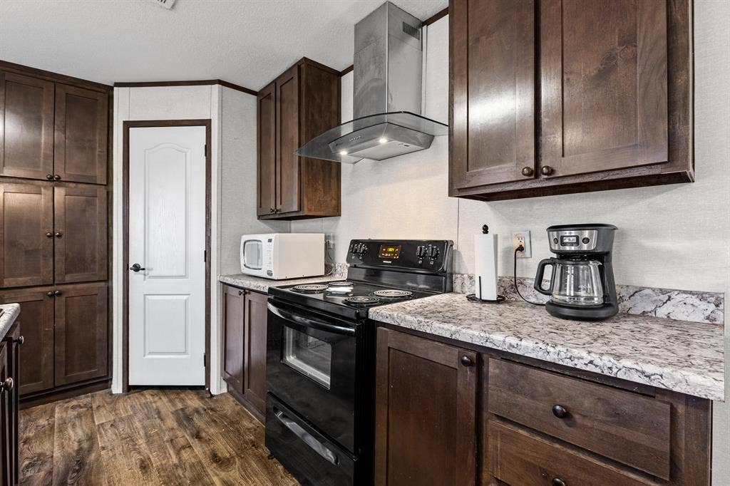 Kitchen featuring rich wood-finish cabinetry, patterned laminate countertops, a stainless steel chimney-style range hood, black range, and wood-finish flooring
