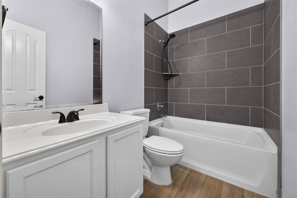 701 Abbey Lane, Midlothian, TX 76065 Bathroom featuring a white vanity cabinet with a solid surface countertop, matte black faucet hardware, wood-finish flooring, and a shower-tub combination with horizontal charcoal-toned tile surround