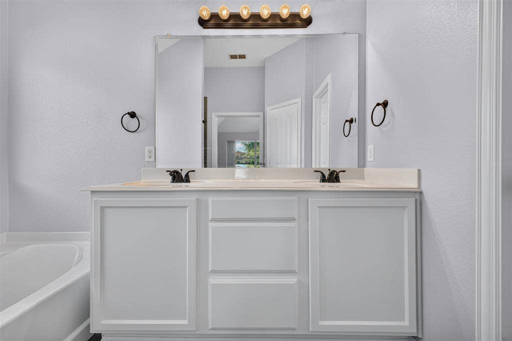 701 Abbey Lane, Midlothian, TX 76065 White dual-sink vanity featuring shaker-style cabinetry, solid-surface countertop, bronze-finish hardware, and a large frameless mirror with five-light vanity fixture