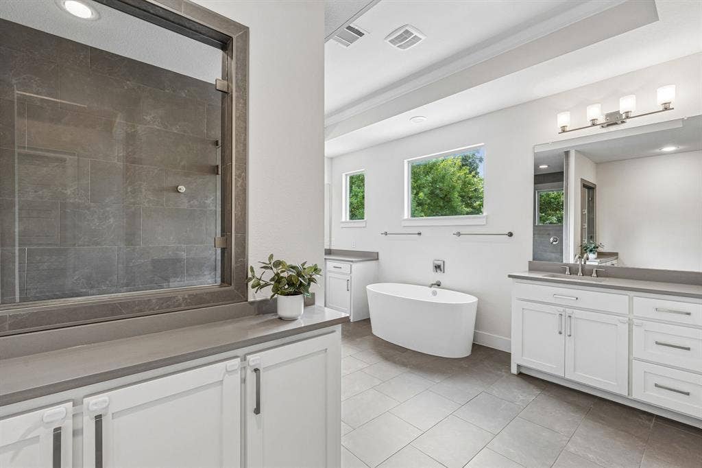 Freestanding soaking tub, double-sink vanity with quartz countertops, large-format tile flooring, and walk-in shower with dark tile surround