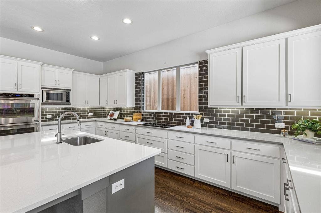 Kitchen featuring a central island with a solid surface countertop, white shaker-style cabinetry, a dark brick-style backsplash, stainless steel built-in appliances, and wood-finish flooring