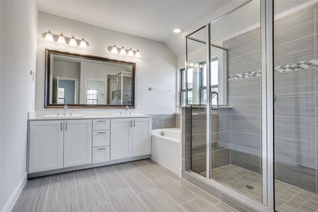 6341 Fortuna Lane, McKinney, TX 75070 Dual-sink vanity with white cabinetry and countertop, framed wall mirror, deep soaking tub, and glass-enclosed shower with horizontal wall tile
