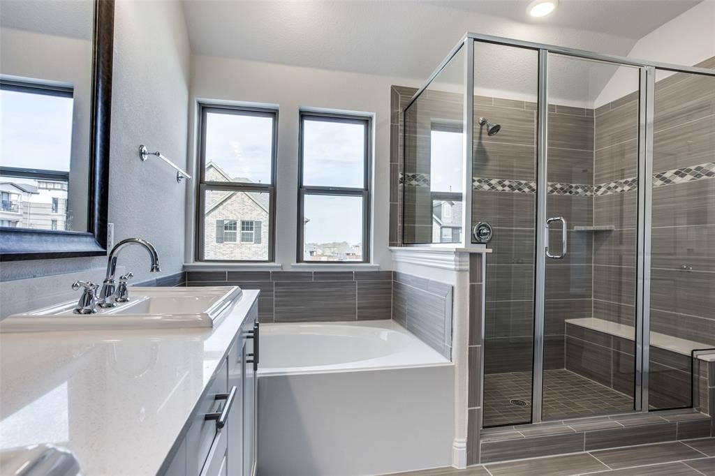 6341 Fortuna Lane, McKinney, TX 75070 Bathroom featuring a dual-sink vanity with white stone-finish countertop, deep soaking tub, and a walk-in glass-enclosed shower with tiled surround, accent inlay, and built-in bench