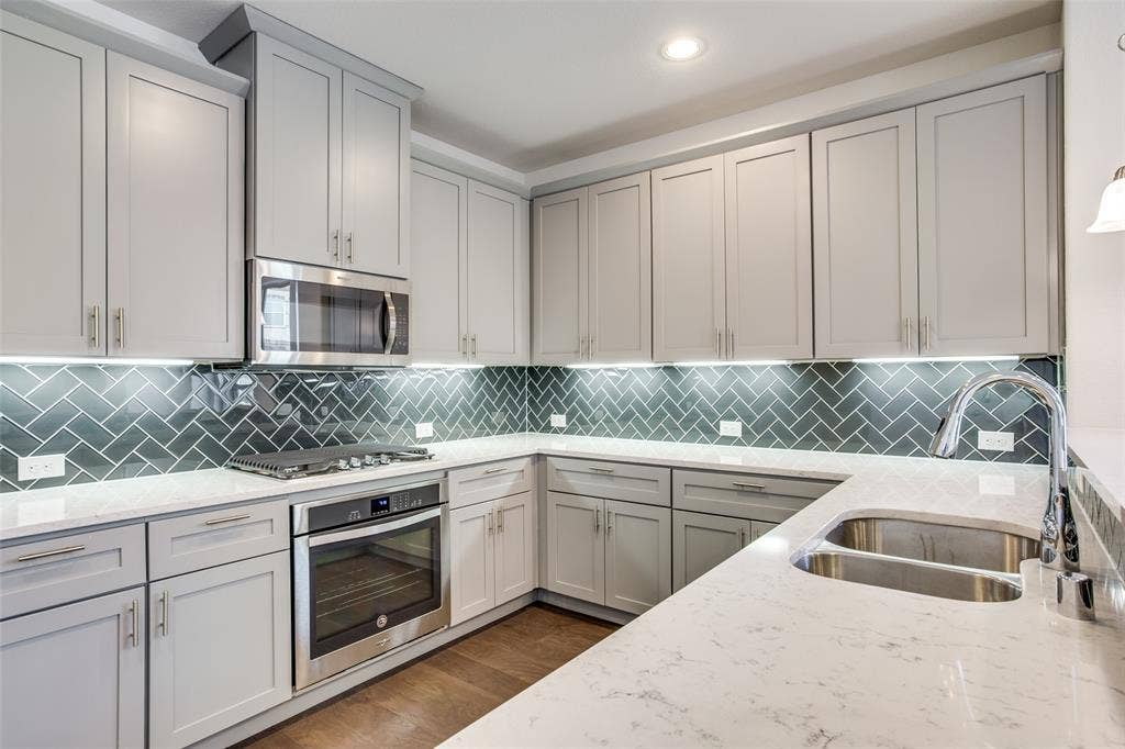 6341 Fortuna Lane, McKinney, TX 75070 Kitchen featuring gray shaker-style cabinetry, white quartz countertops, a herringbone-patterned tile backsplash, stainless steel built-in appliances, and a dual-basin undermount sink