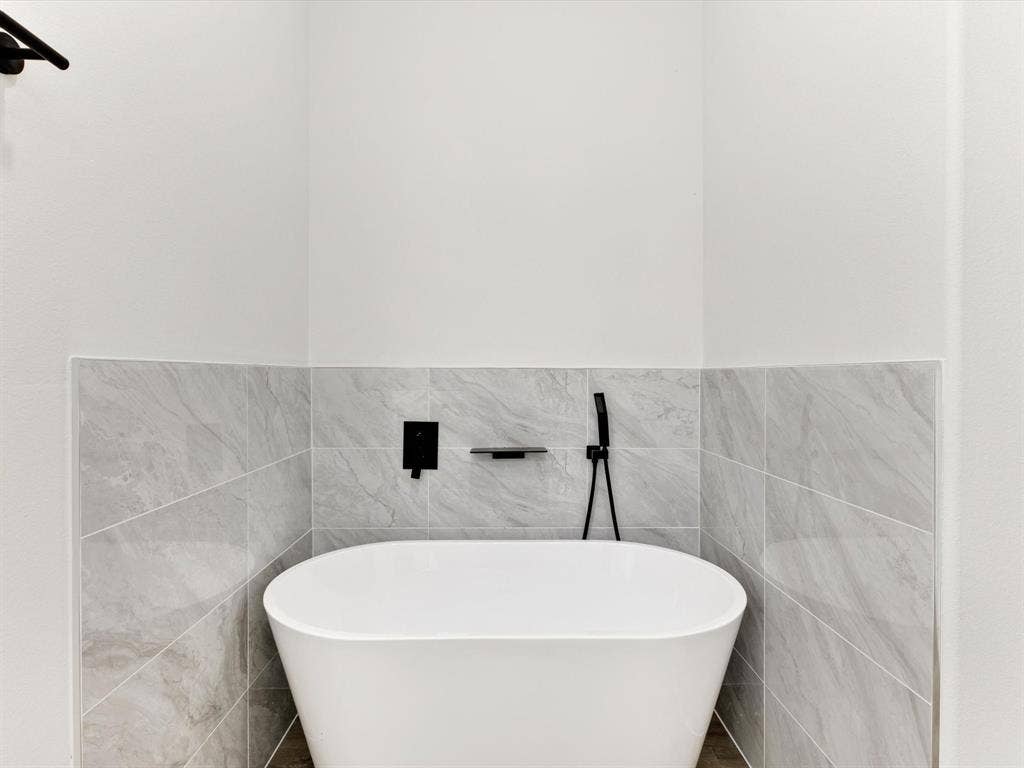 475 Loren Boyd Road, Combine, TX 75159 Freestanding white soaking tub with matte black wall-mounted faucet, handheld shower wand, built-in soap niche, and grey stone-finish tile surround