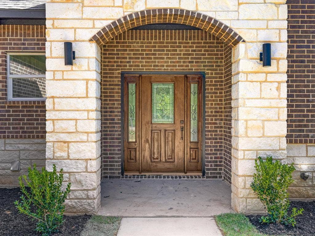 475 Loren Boyd Road, Combine, TX 75159 Entryway featuring a arched stone portico, brick facade, custom wood door with decorative glass paneling, dual sidelights, and exterior wall-mounted lighting