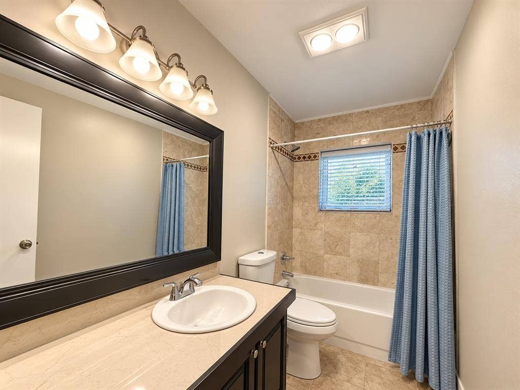 7525 Holly Hill Drive, #3, Dallas, TX 75231 Bathroom featuring dark wood-finish cabinetry, a neutral countertop with an integrated basin, a large framed vanity mirror, and stone-tile tub surround with decorative inlay