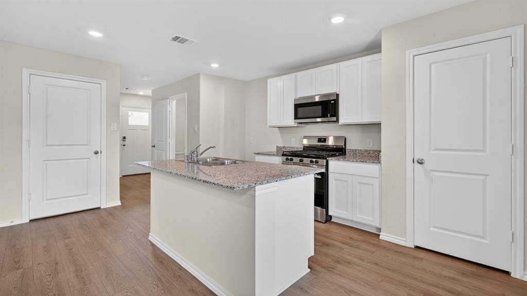 Kitchen featuring a granite-topped island with an undermount sink, stainless steel appliances, white cabinetry, and wood-finish flooring