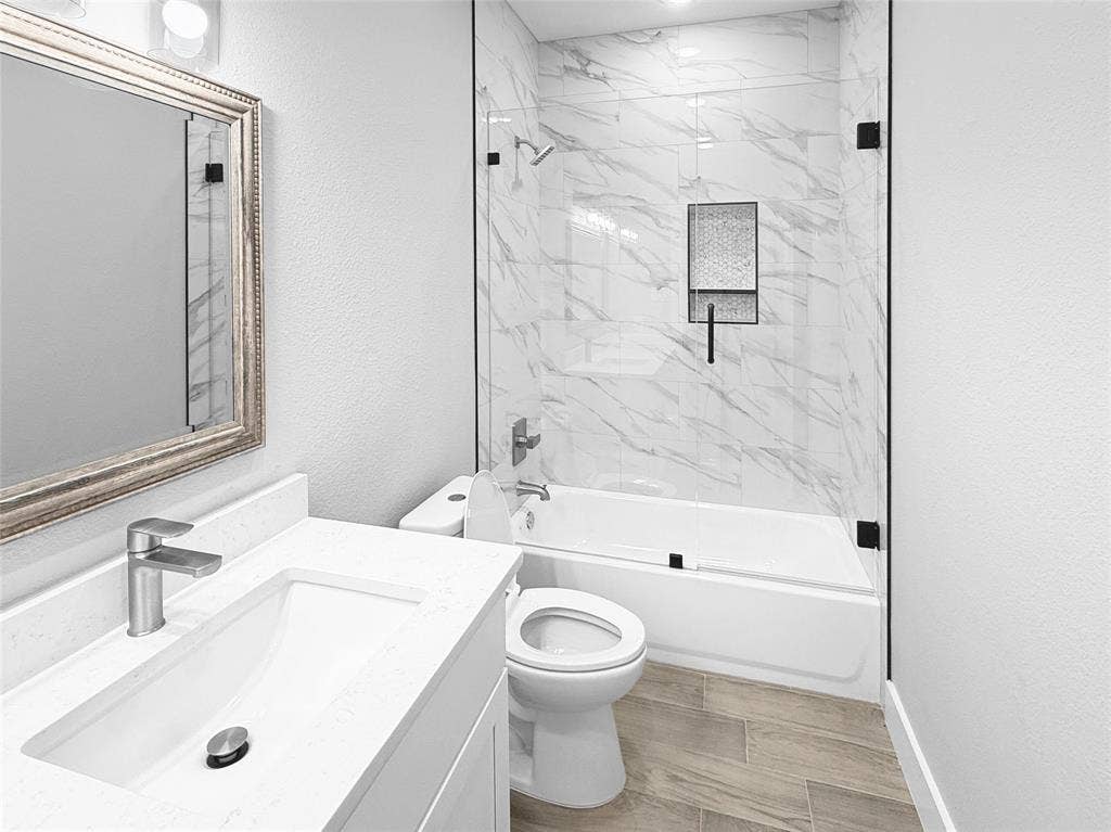 3025 Red Oak Drive, Granbury, TX 76048 Bathroom featuring a vanity with an integrated rectangular sink and stone-finish countertop, wood-finish flooring, and a tub-shower combination with floor-to-ceiling tile surround, glass enclosure, and built-in recessed shelving