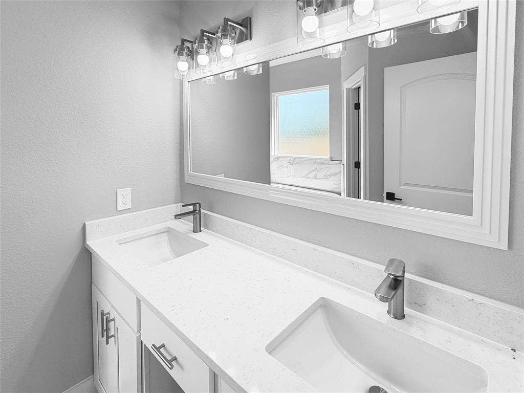 3025 Red Oak Drive, Granbury, TX 76048 Dual-sink vanity featuring a white composite countertop, undermount basins, brushed nickel hardware, and a framed mirror with integrated multi-bulb lighting