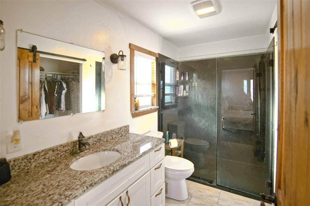 325 & 321 Grassy Way, Elm Mott, TX 76640 Stone-topped vanity featuring white cabinetry, glass-enclosed shower with dark metal hardware, wall-mounted light fixtures, and wood-finish trim