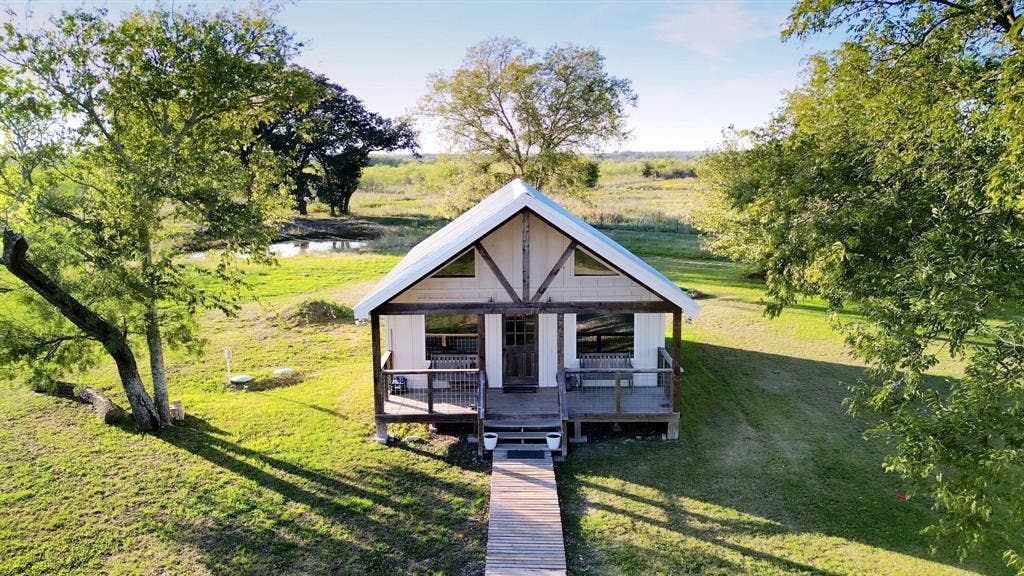325 & 321 Grassy Way, Elm Mott, TX 76640 Gable-roof cottage featuring a covered front porch, timber-framed exterior accents, wood-plank walkway, and expansive grounds with mature trees