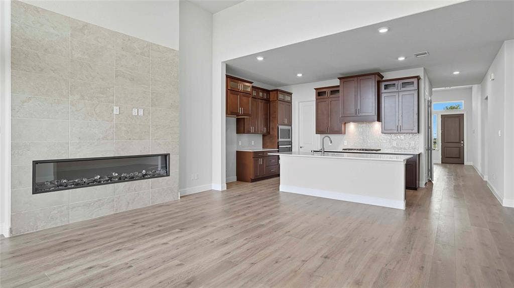 Fireplace with stone-finish surround, wood-finish flooring, recessed lighting, and kitchen featuring dark-tone cabinetry with stone-finish countertops