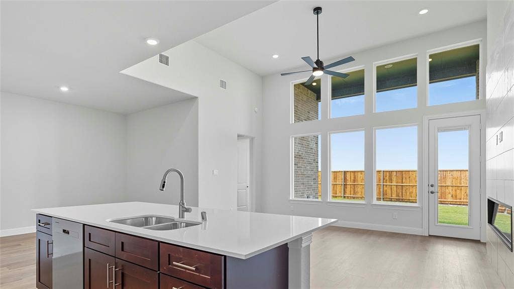 Expansive interior featuring high ceilings, a kitchen island with stone countertops and a dual-basin stainless steel sink, large windows, wood-finish flooring, and a sleek ceiling fan