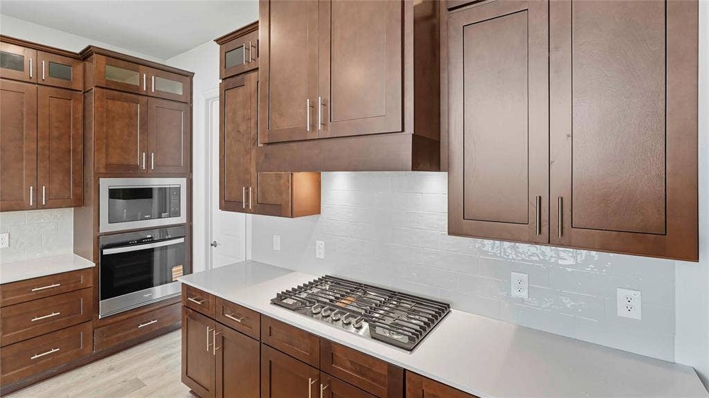 Kitchen featuring dark-finish cabinetry, light-toned stone-surface countertops, a tile backsplash, a built-in stainless steel double oven, and a gas cooktop