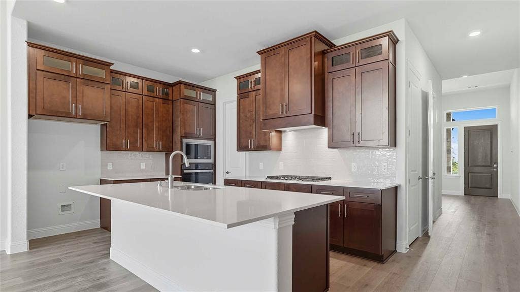 Kitchen featuring cabinetry with glass-paneled upper doors, expansive quartz countertops, an integrated cooktop, built-in wall oven, and wood-finish flooring