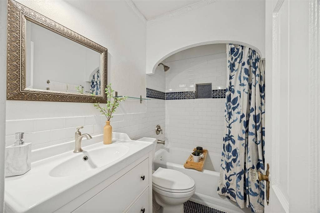 7011 Hammond Avenue, Dallas, TX 75223 White vanity featuring an integrated sink with a metallic faucet, wall-mounted mirror with a textured metallic frame, tub-shower combination with white subway tile surround, decorative accent tile border, and an arched architectural opening