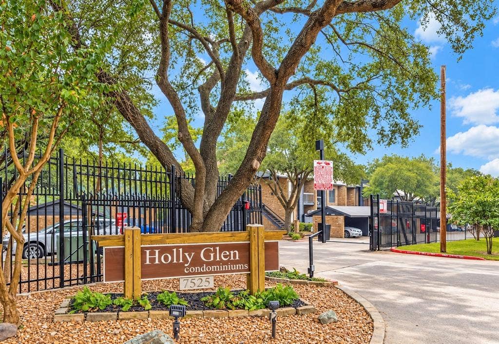 7525 Holly Hill Drive, #20, Dallas, TX 75231 Gated community entrance featuring metal perimeter fencing, a stone-bordered garden bed, mature tree canopy, and covered parking structures