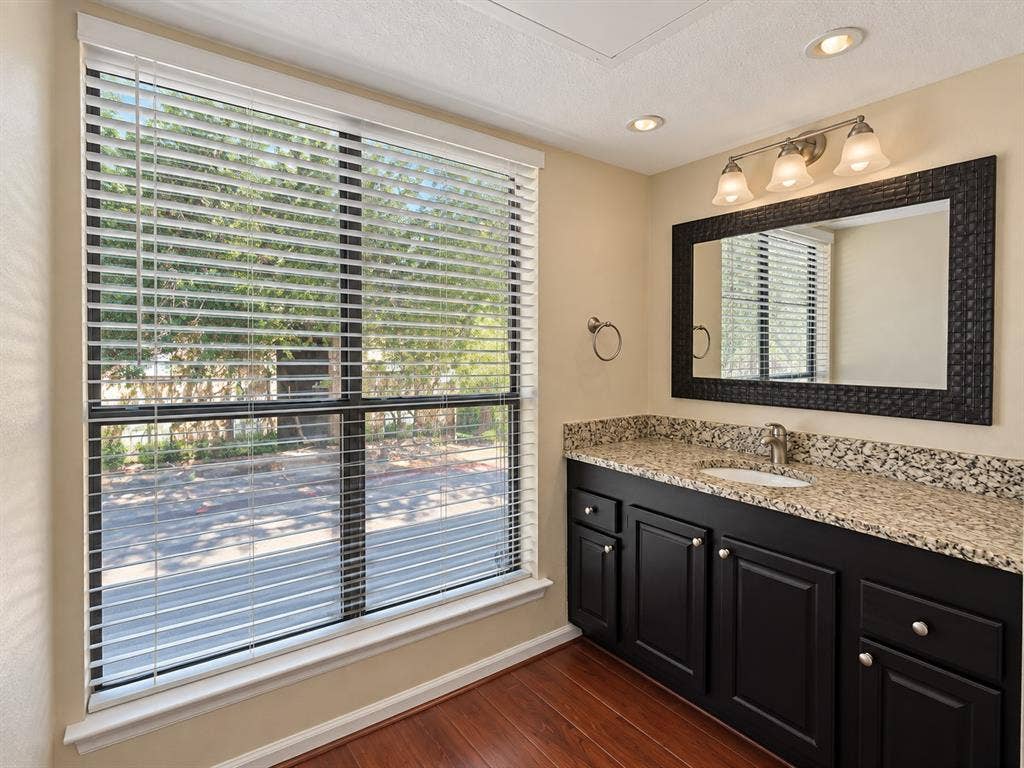 7525 Holly Hill Drive, #20, Dallas, TX 75231 Bathroom featuring wood-finish flooring, a vanity with dark cabinetry and a stone-style countertop, an integrated sink, a framed wall mirror, and large windows with horizontal blinds