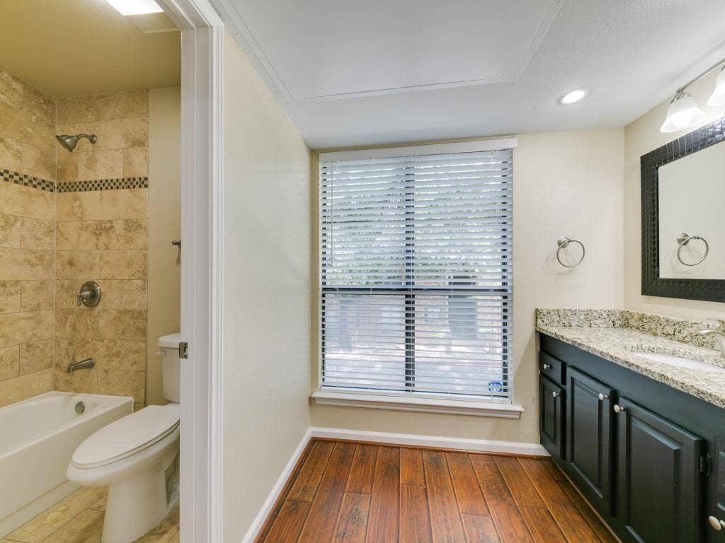 7525 Holly Hill Drive, #20, Dallas, TX 75231 Bathroom featuring wood-finish flooring, dark-finish cabinetry with a stone-composite countertop, and a shower-tub combination with tile surround and a decorative mosaic inlay