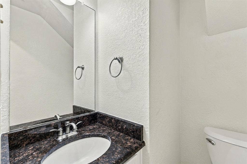 6821 Douglas Creek, Plano, TX 75023 Dark stone-finish countertop with an integrated oval under-mount sink, chrome dual-handle faucet, large wall-mounted mirror, and circular chrome towel ring