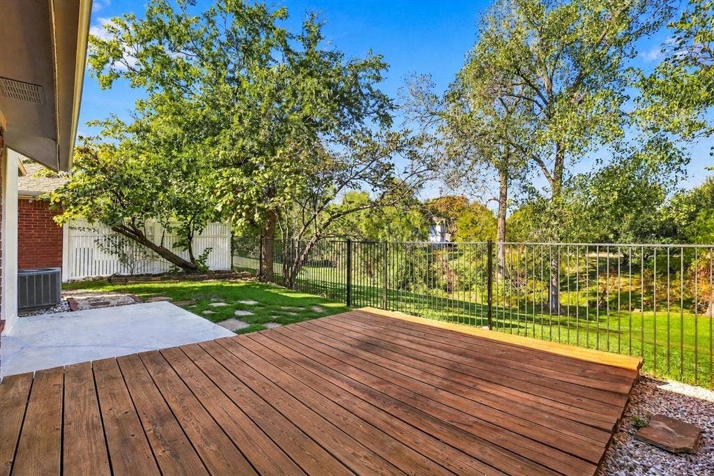 6821 Douglas Creek, Plano, TX 75023 Backyard featuring a raised wooden deck, concrete patio area, black metal perimeter fencing, and mature trees