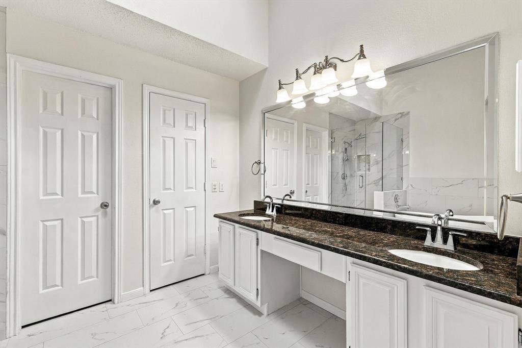 6821 Douglas Creek, Plano, TX 75023 Dual-sink vanity with dark stone countertop, white shaker-style cabinetry, large beveled-edge mirror, and five-light vanity fixture
