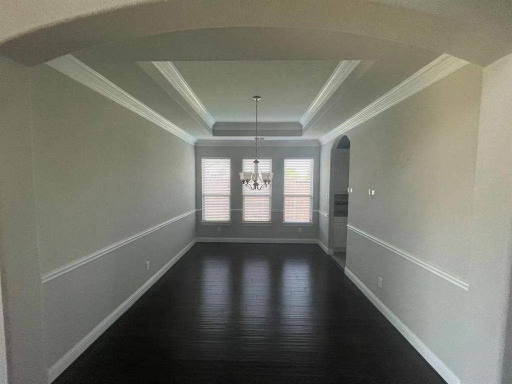 15112 Morning Star Lane, Frisco, TX 75035 Interior space featuring dark wood-finish flooring, a tray ceiling with crown molding, chair rail trim, and three windows with neutral blinds beneath a central pendant light fixture