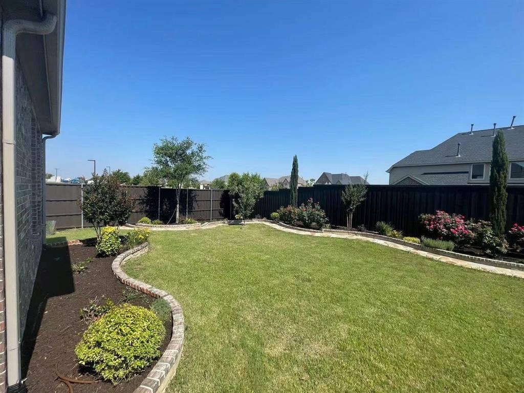 15112 Morning Star Lane, Frisco, TX 75035 Backyard featuring a manicured lawn, stone-bordered garden beds, dark wood-style perimeter fencing, and mature ornamental shrubs