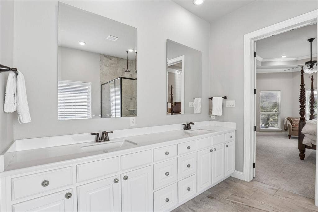 676 Princeton Way, Rockwall, TX 75087 Primary bath featuring dual-sink vanity with white cabinetry, light-toned stone-style countertop, rectangular undermount sinks, and dark metal hardware paired with wood-finish tile flooring