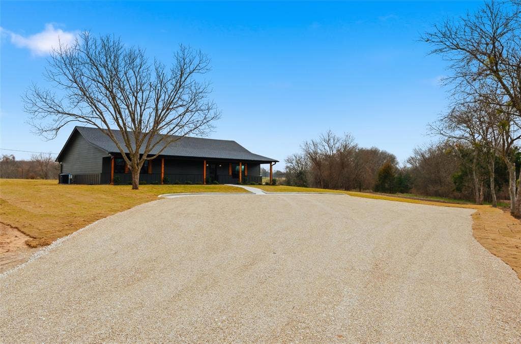 2303 Pilgrim Lane, Lorena, TX 76655 Single-story exterior featuring dark siding, a covered front porch with wooden support posts, a concrete walkway, and a gravel driveway situated on expansive acreage