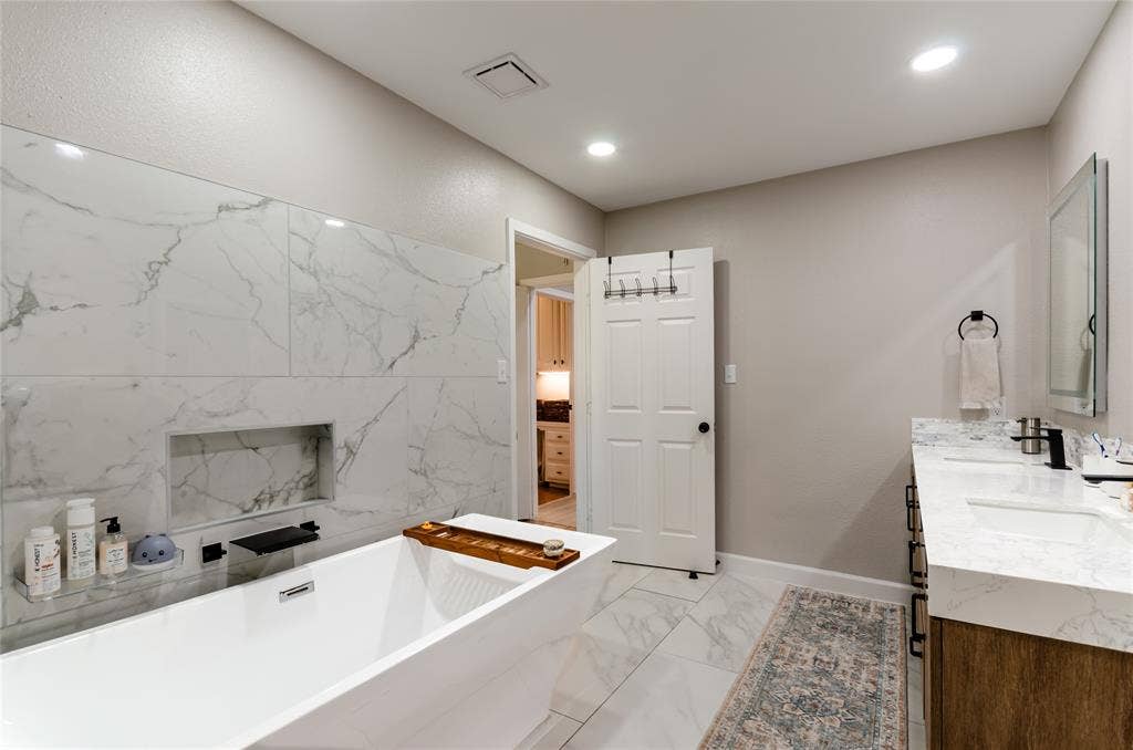 Freestanding soaking tub with a built-in wall niche, dual sink vanity with stone countertops, and recessed lighting