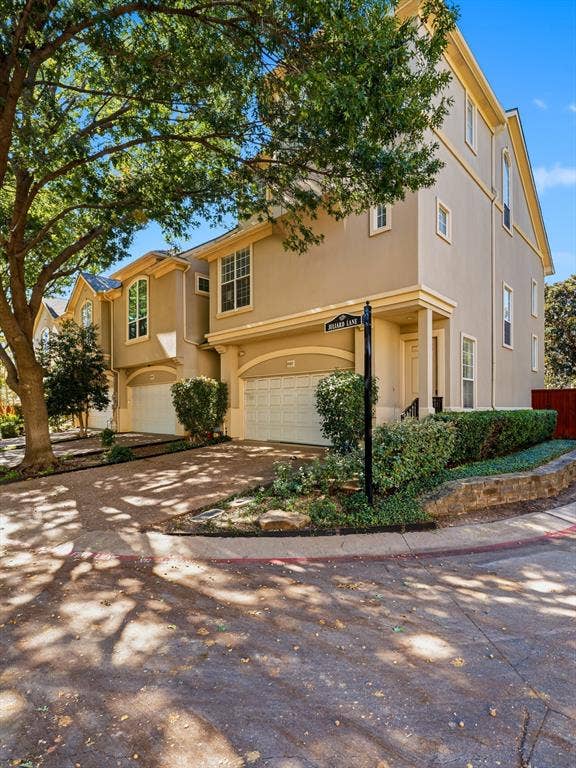 14567 Juliard Lane, Addison, TX 75001 Multi-story exterior featuring a neutral stucco facade, arched garage entries, a textured aggregate driveway, and tiered stone retaining walls