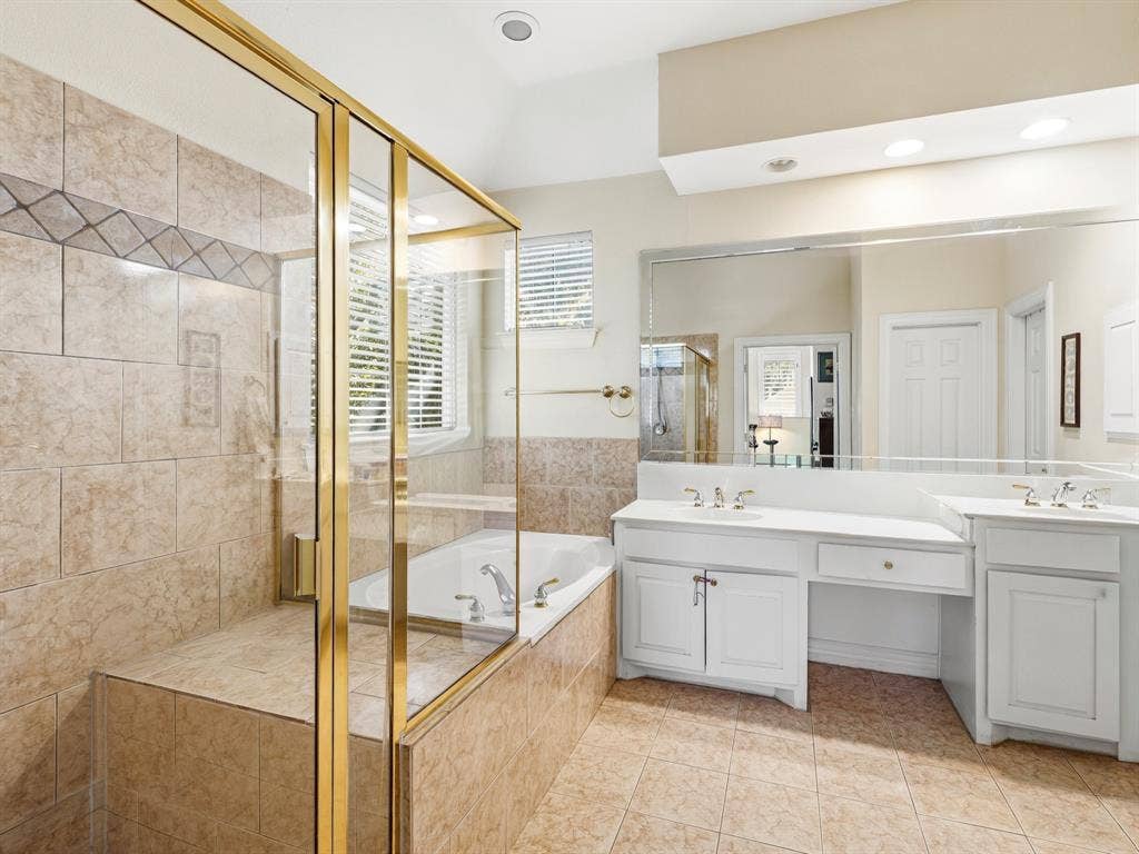 14567 Juliard Lane, Addison, TX 75001 Bathroom featuring a dual-sink vanity with white cabinetry, a glass-enclosed shower with tiled walls and gold-tone trim, and a deep soaking tub