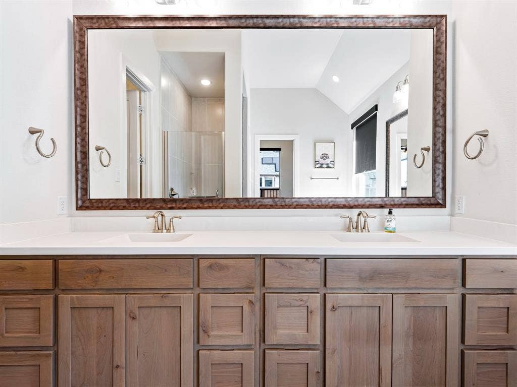 Dual-sink vanity with a solid-surface countertop, wood-finish shaker cabinetry, large framed mirror, and dual brushed-finish faucets
